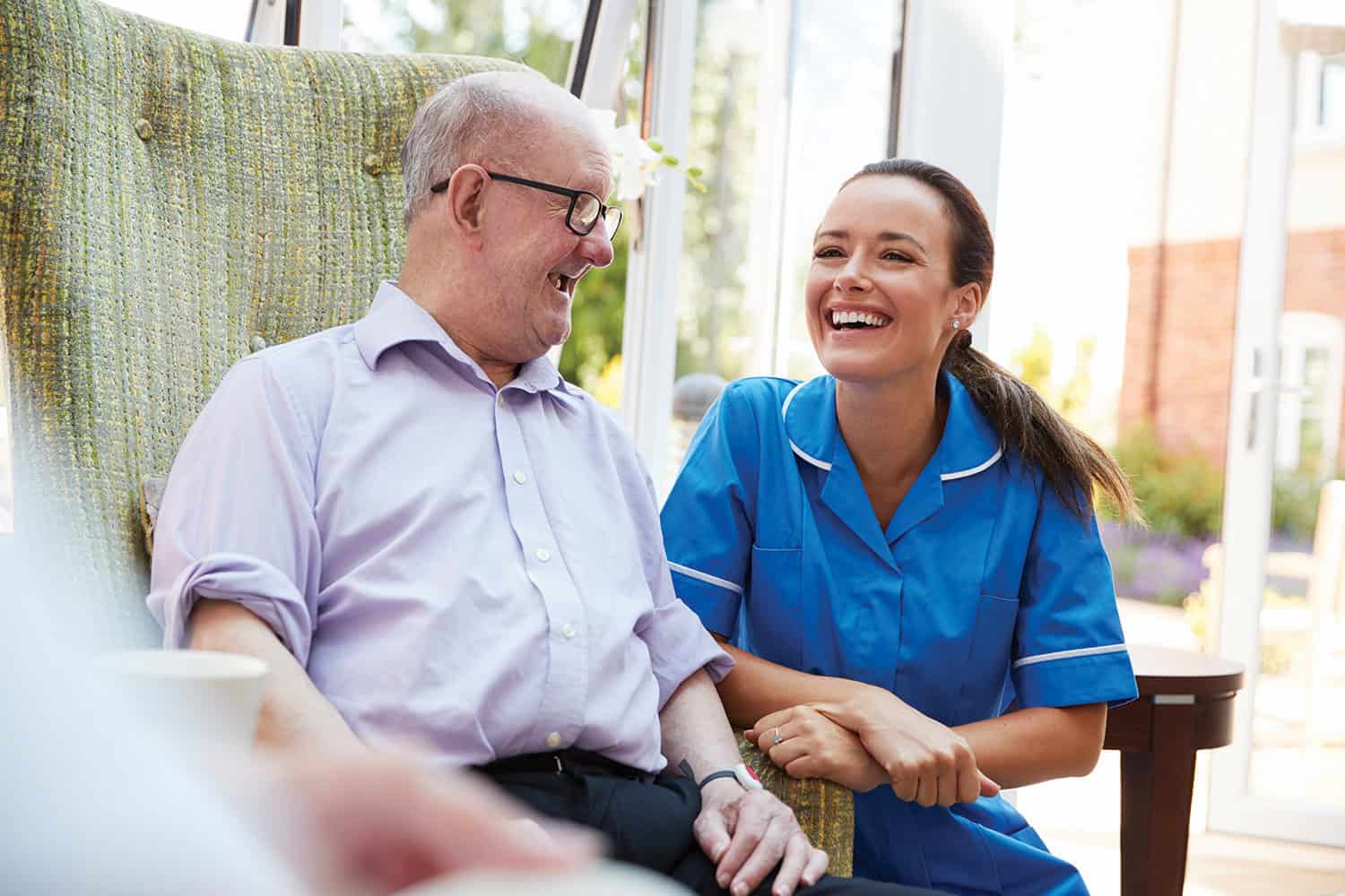 photo of older man in glasses in armchair, laughing with younger female carer