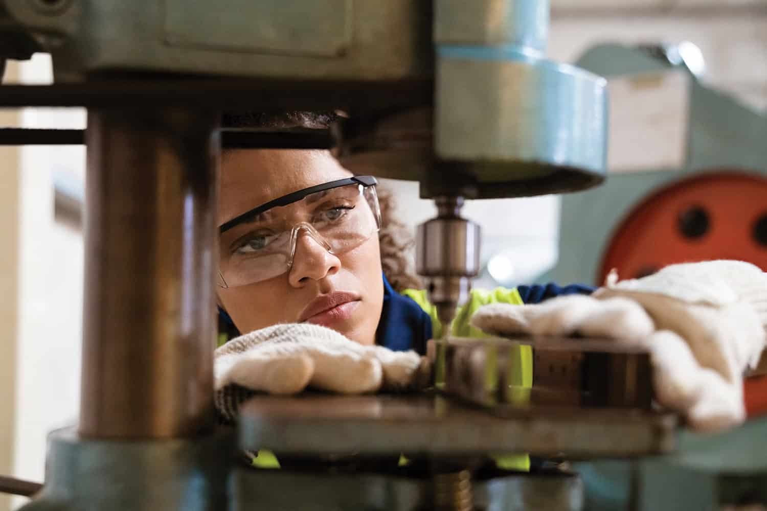photo of close-up of female apprentice wearing safety glasses and gloves, and looking through a fixed drill point; photo of blurred vehicles on a busy stretch of motorway at sunset