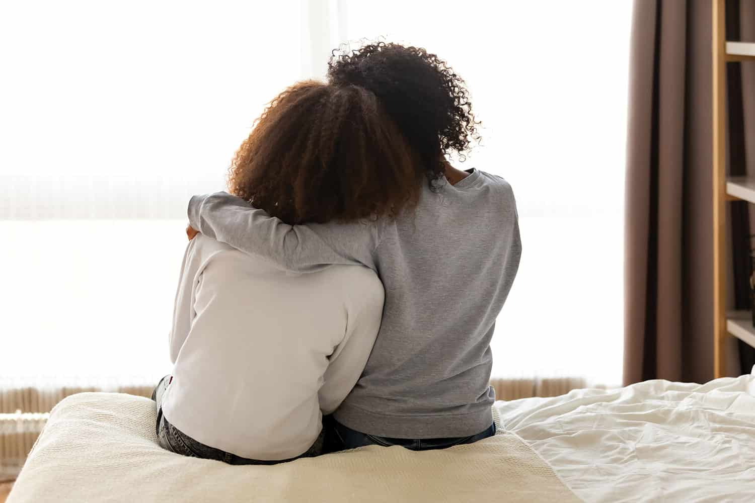 photo from behind of older woman with arm around younger woman, both sitting on a bed