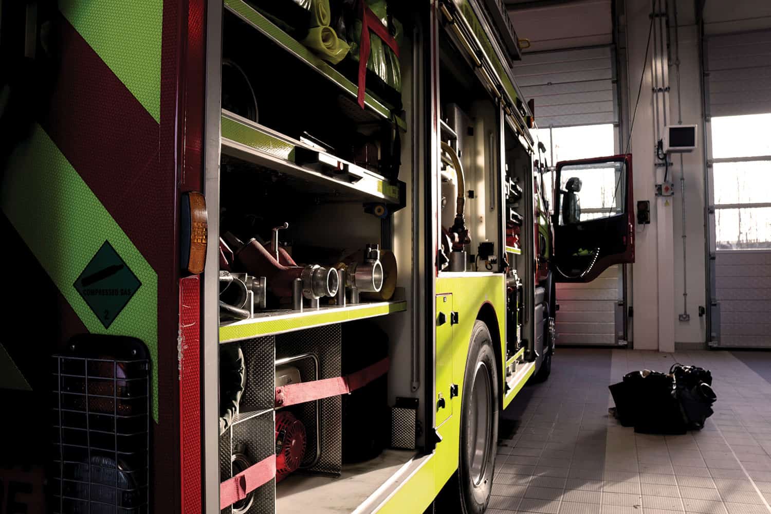 : photo of fire truck with side compartments open to display hose and other fire fighting equipment, inside the bay of a fire station