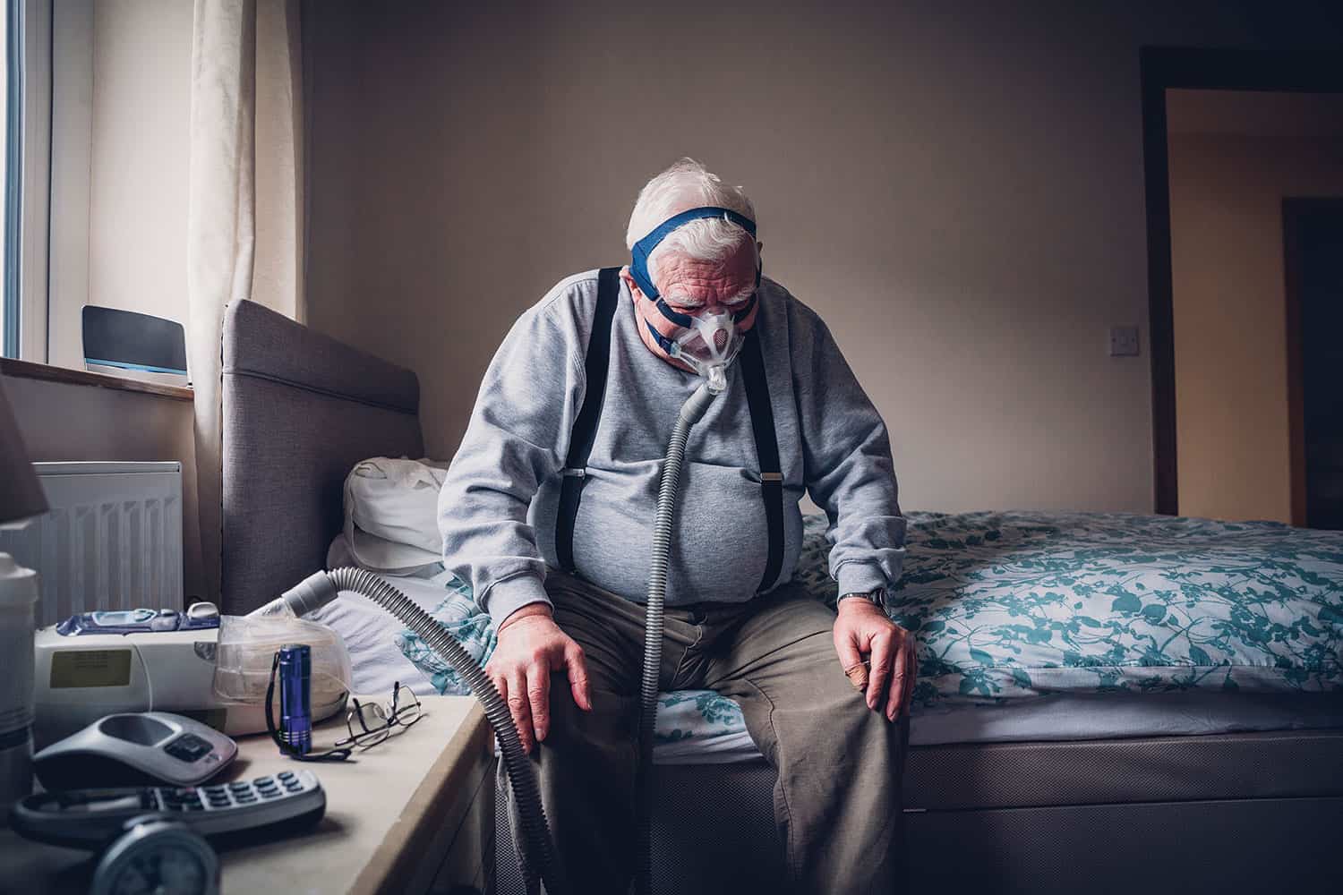 Older overweight man sitting on side of bed at home, with a face mask strapped over his face connected to a breathing machine.
