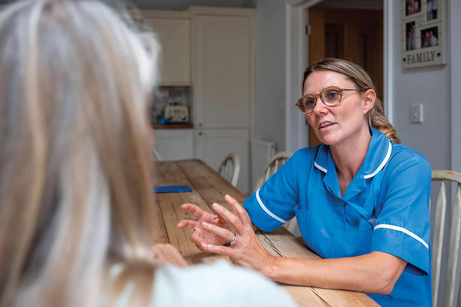 Female health visitor in blue uniform talking to client across kitchen table – pictured from behind the client.