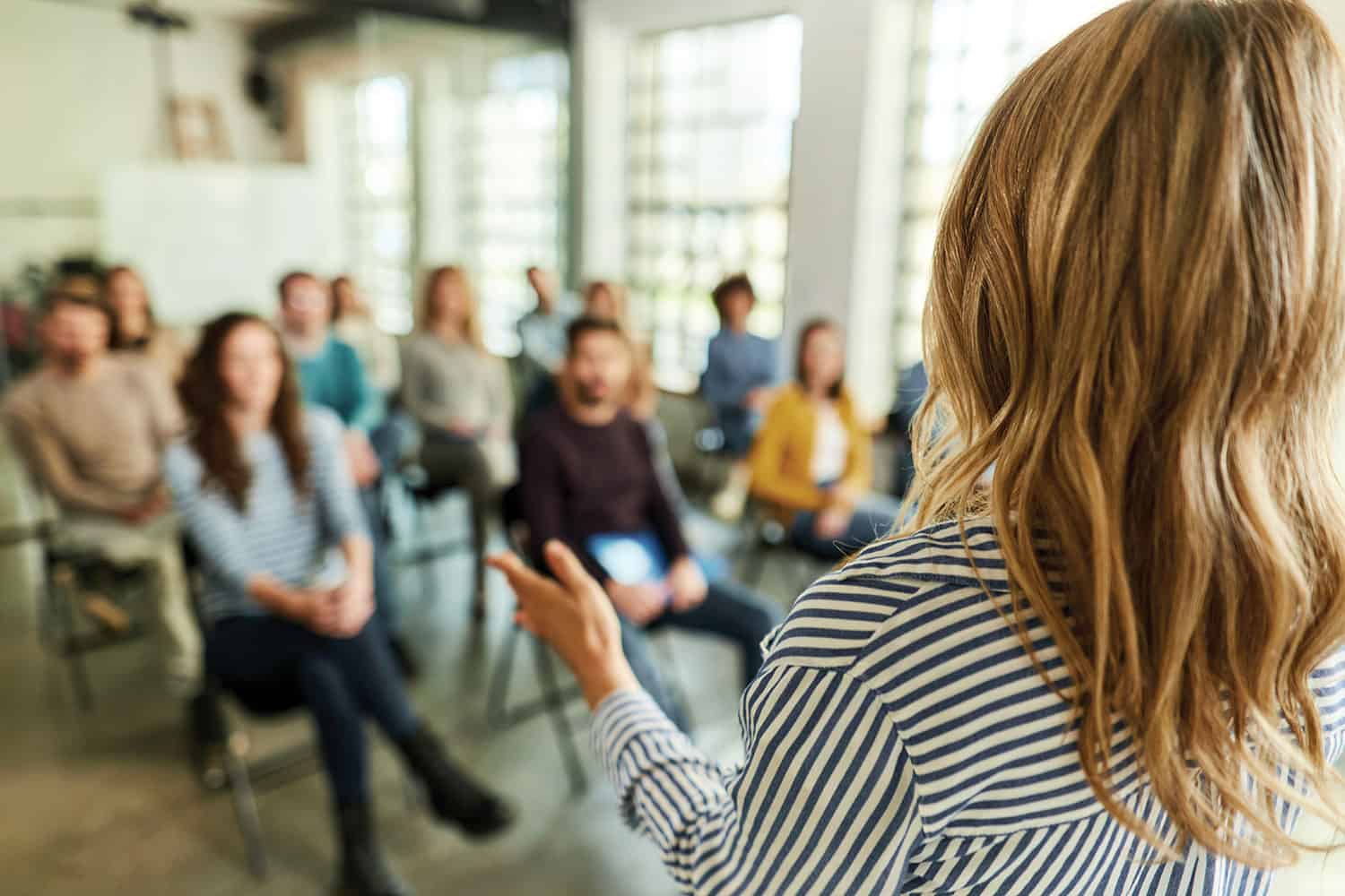 young woman standing with back to camera talking to blurred group of people sitting on chairs