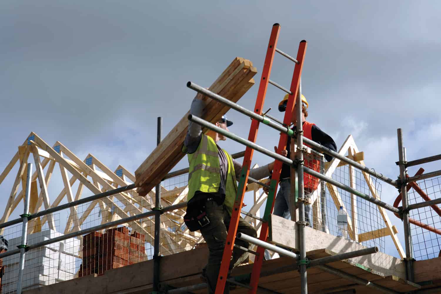 photo of workman carrying timbers on shoulder along scaffolding walkway alongside roof being built on new house)