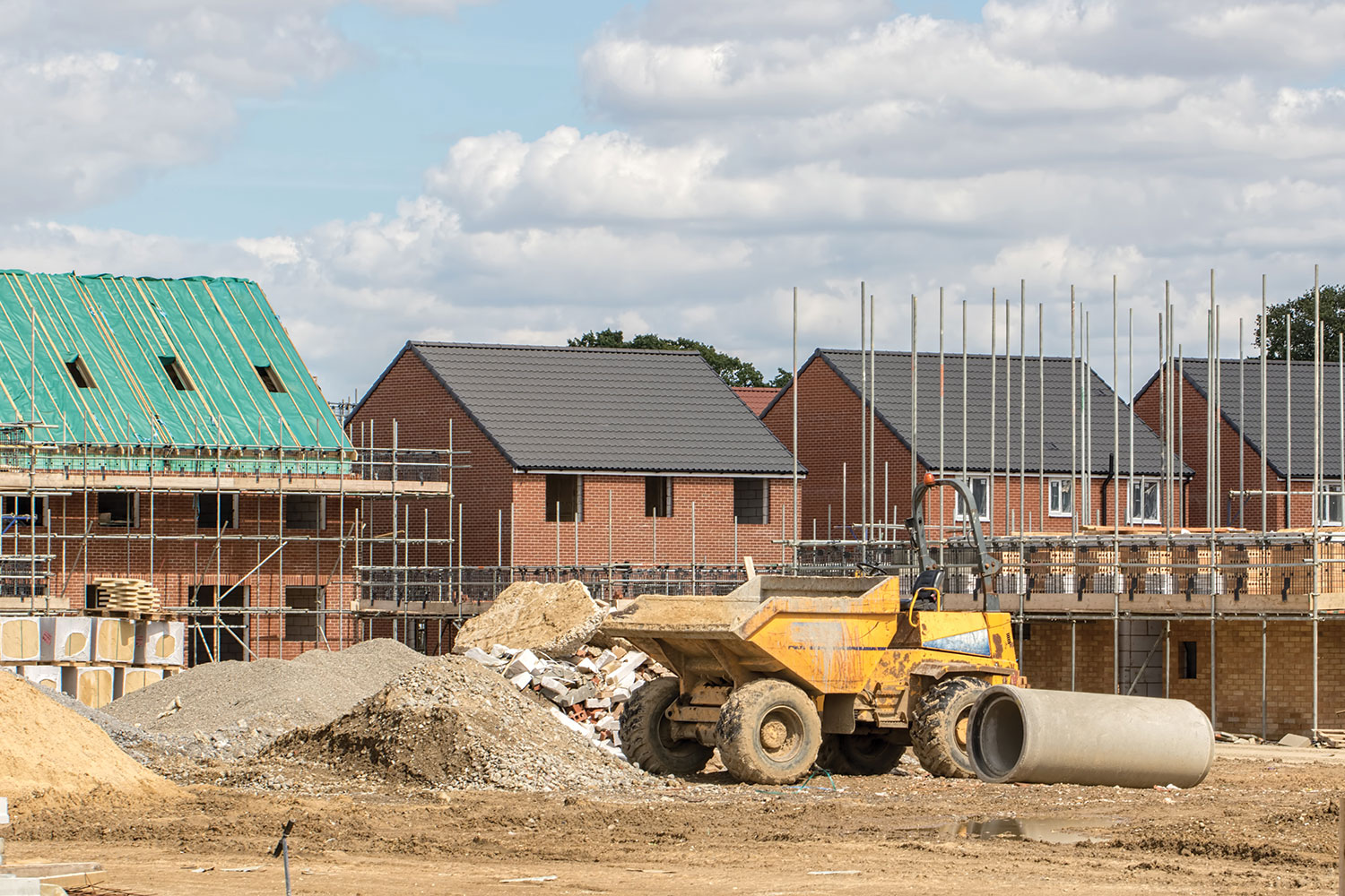 new houses being built with scaffolding around them and some complete, with yellow dumper truck in foreground between piles of sand, gravel and other building materials