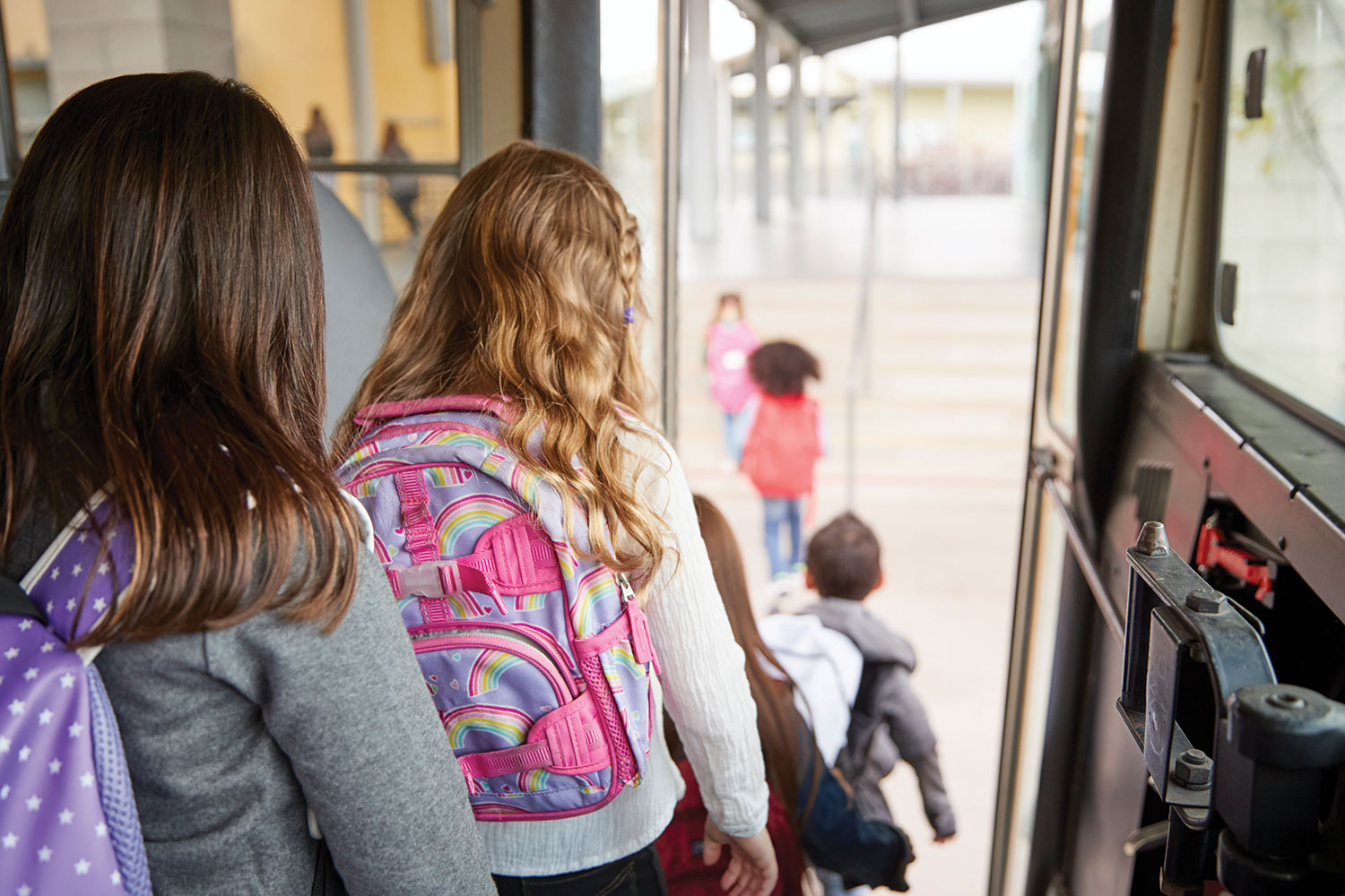view from behind of primary school children with colourful backpacks disembarking from a school bus
