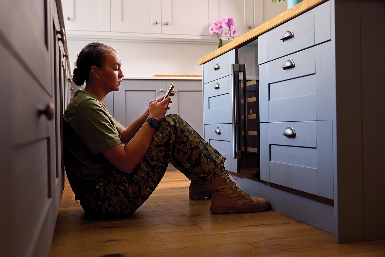woman in army combat fatigues sitting on floor of kitchen texting on her mobile phone