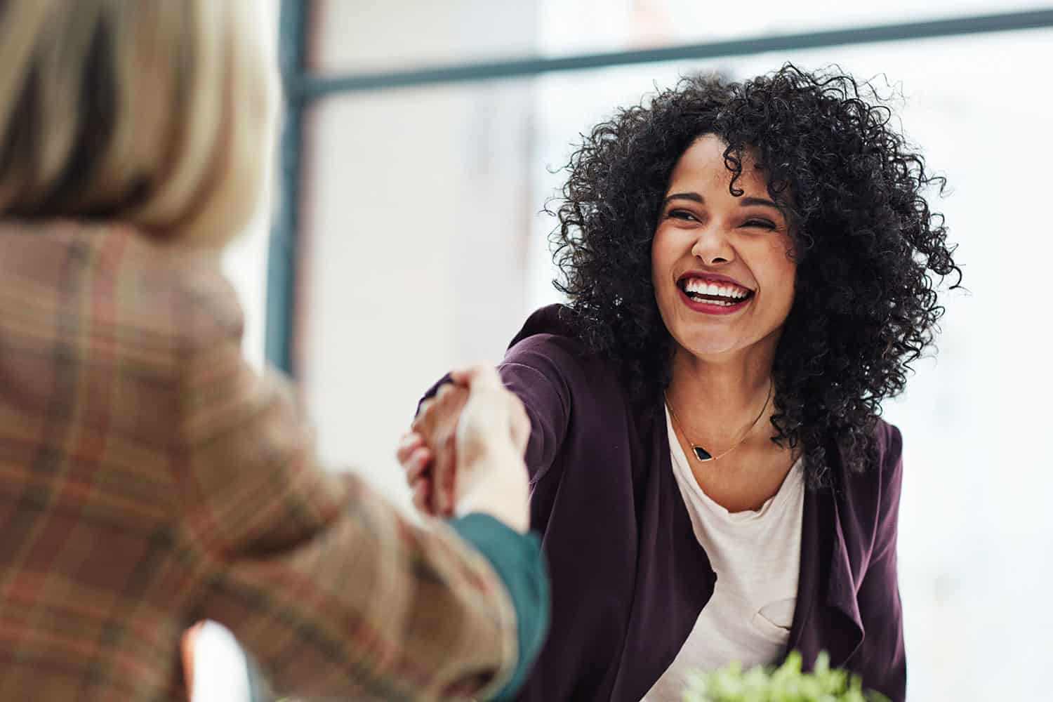 : from behind, woman in suit shaking hands with smiling younger woman who has been offered a job